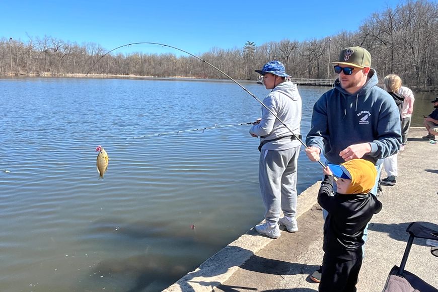 A volunteer helps a young child land a panfish while fishing from a pier.