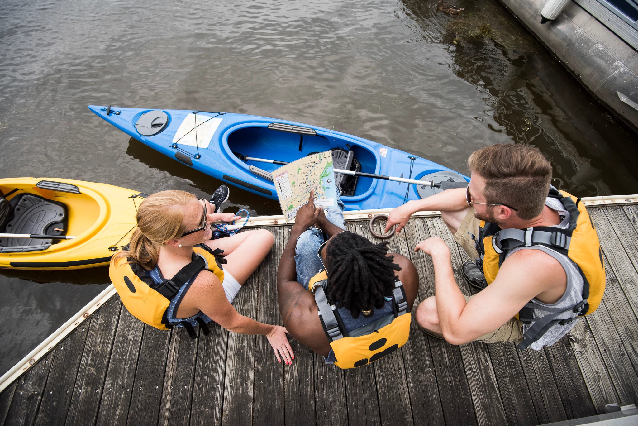 Three people kneel on a dock next to kayaks in the water looking at a map. They are all wearing life jackets.