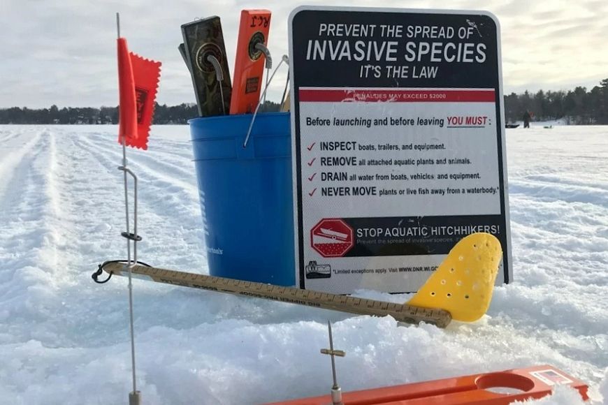A sign titled "Prevent The Spread Of Invasive Species - It's The Law" propped against a 5-gallon bucket filled with ice fishing gear on a frozen lake. 