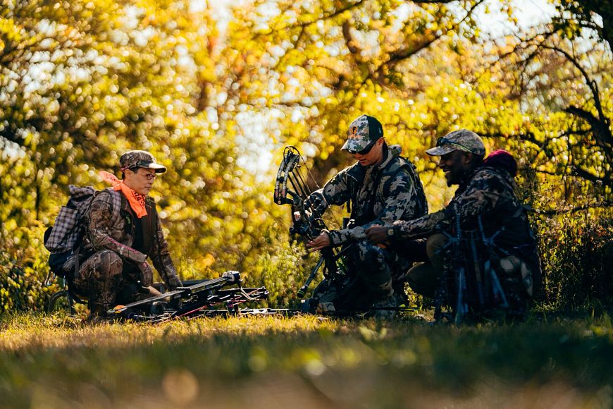 three hunters holding hunting equipment crouching in a wooded area
