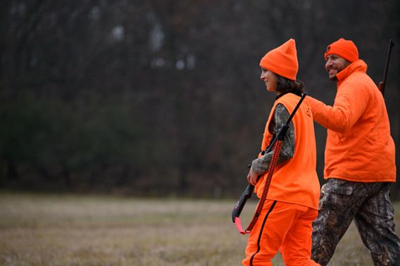 A man in an orange coat and hat and camo pants mentors a woman in blaze orange and camo, holding a riffle.