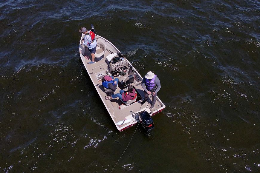 An aerial view of a boat on the water, with four individuals wearing life jackets and fishing seen standing on it.
