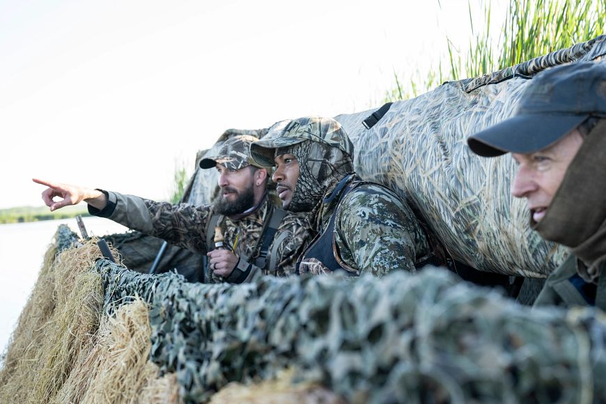 Three people in camouflage clothing are in a duck boat on the edge of a lake, and one person is pointing while holding a duck call.