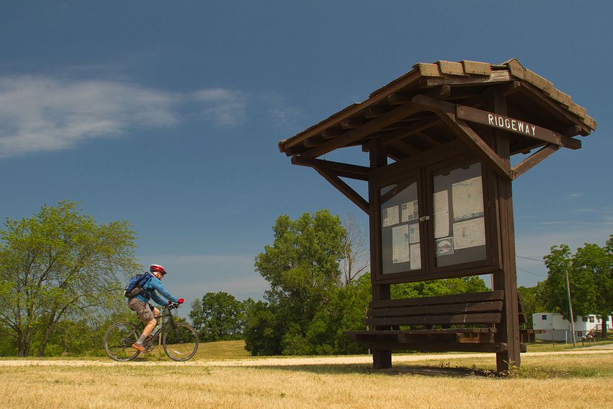 A cyclist biking on the Military Ridge Trail near a trailhead sign on a summer day.