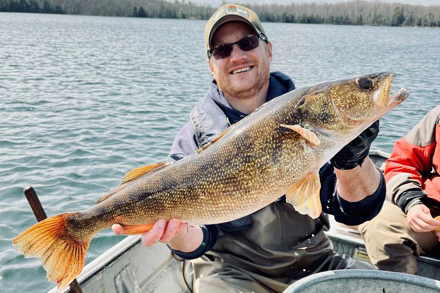 A man in a baseball cap and an inflatable life jacket holds up a large walleye on a small boat.