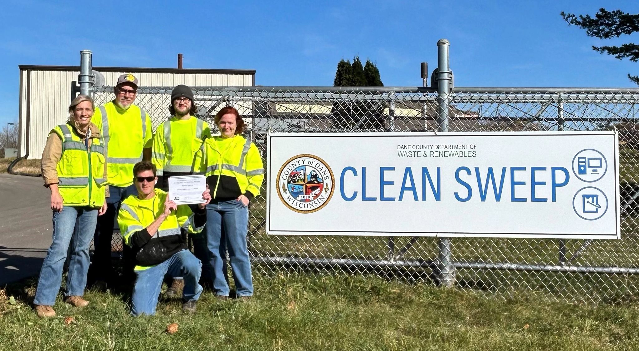 Employees from Dane County Clean Sweep stand outside next to a large sign for the facility while holding their award. All five employees are wearing bright green protective jackets.