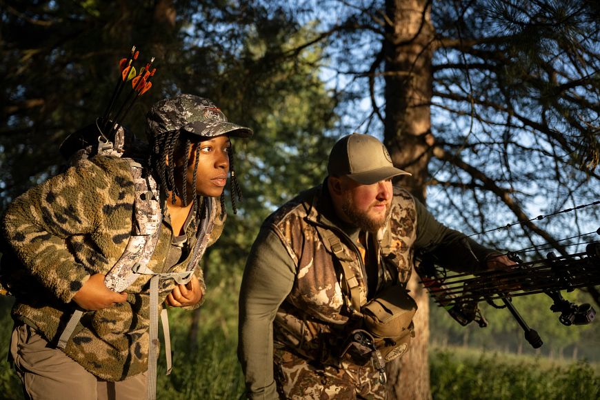 a male and female wearing camouflage carrying bows and arrows looking at something in the forest
