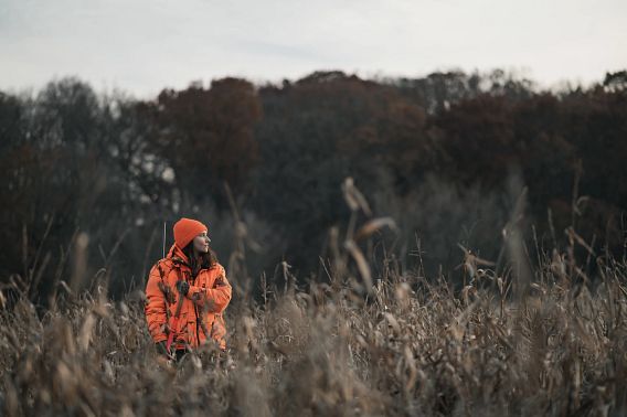 A young woman wearing blaze orange stands in a dried cornfield while she holds a rifle by her side.