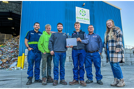Six Green Circle Recycling employees pose outside their facility with their award. In the background is a large pile of collected materials inside their facility.