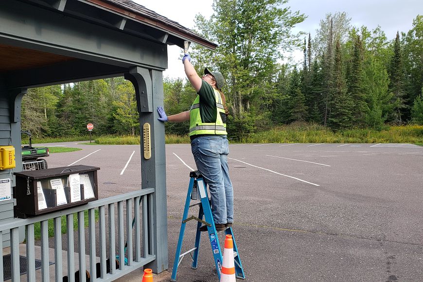 A person wearing a fluorescent vest stands on a ladder painting the awning of a wooden building.