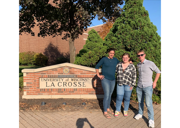 Three University of Wisconsin-La Crosse representatives stand outside next to a brick and stone sign that has the school's name.