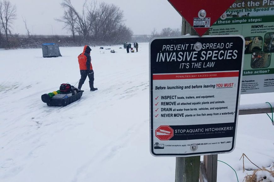 A sign that reads "Prevent the spread of invasive species, it's the law" is seen in the foreground with people ice fishing on a snow-cover lake in the background.