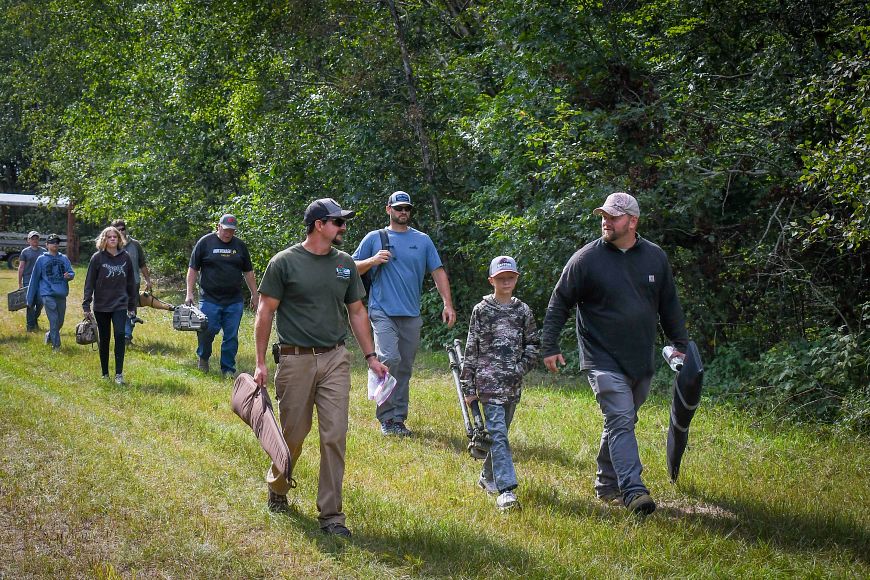 Group of individuals walking through a grassy area with trees, some carrying outdoor gear or gun cases.