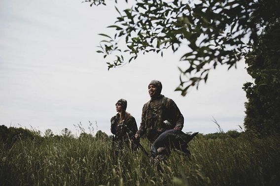 Two hunters, both in camouflage clothing, standing in a field of tall grass. One holds a turkey decoy.
