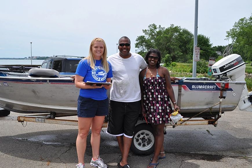 Three adults smiling and standing in front of a boat on a trailer by a lake on a sunny day.