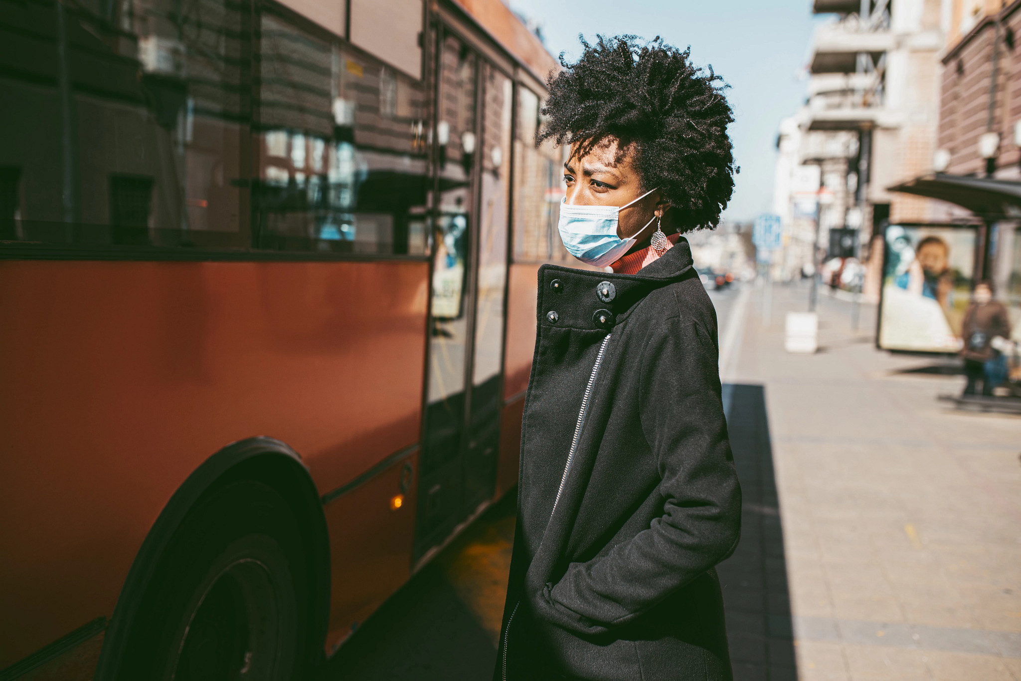 Young African American Woman Standing On City Street Facing A Bus With Protective Mask On Her Face. 