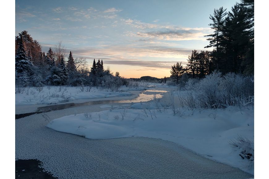 A partially frozen stream winds through a snowy forest landscape under a bright, partially-cloudy sky.