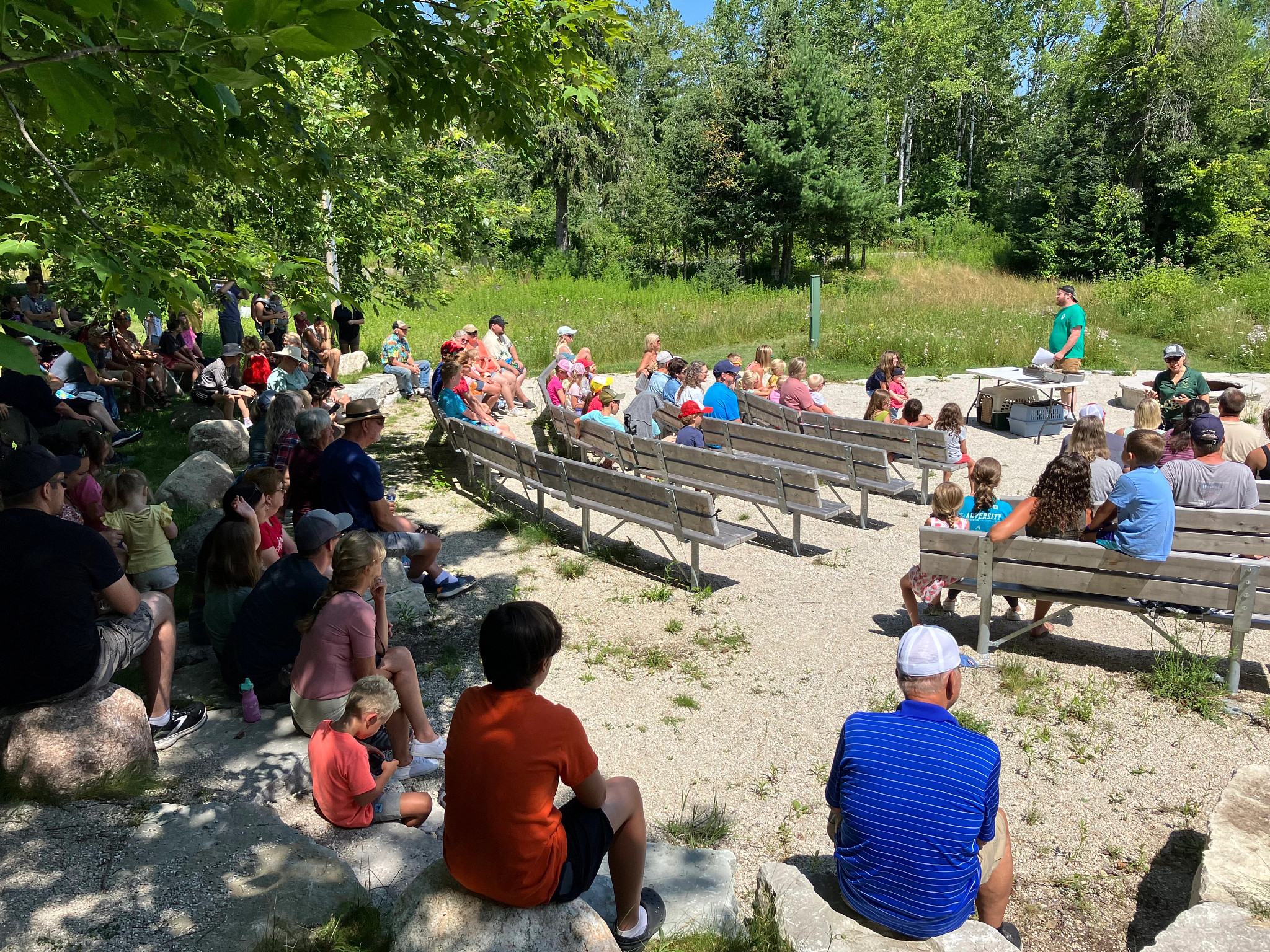 People gather around the amphitheater to see a presentation on a summer day.