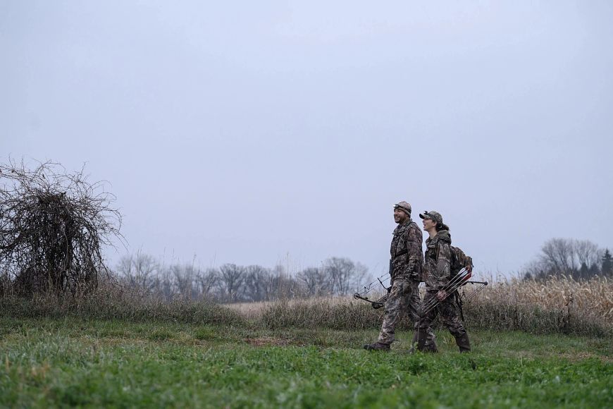 a man and woman in full camouflage walking holding bows in a grassy area against a field