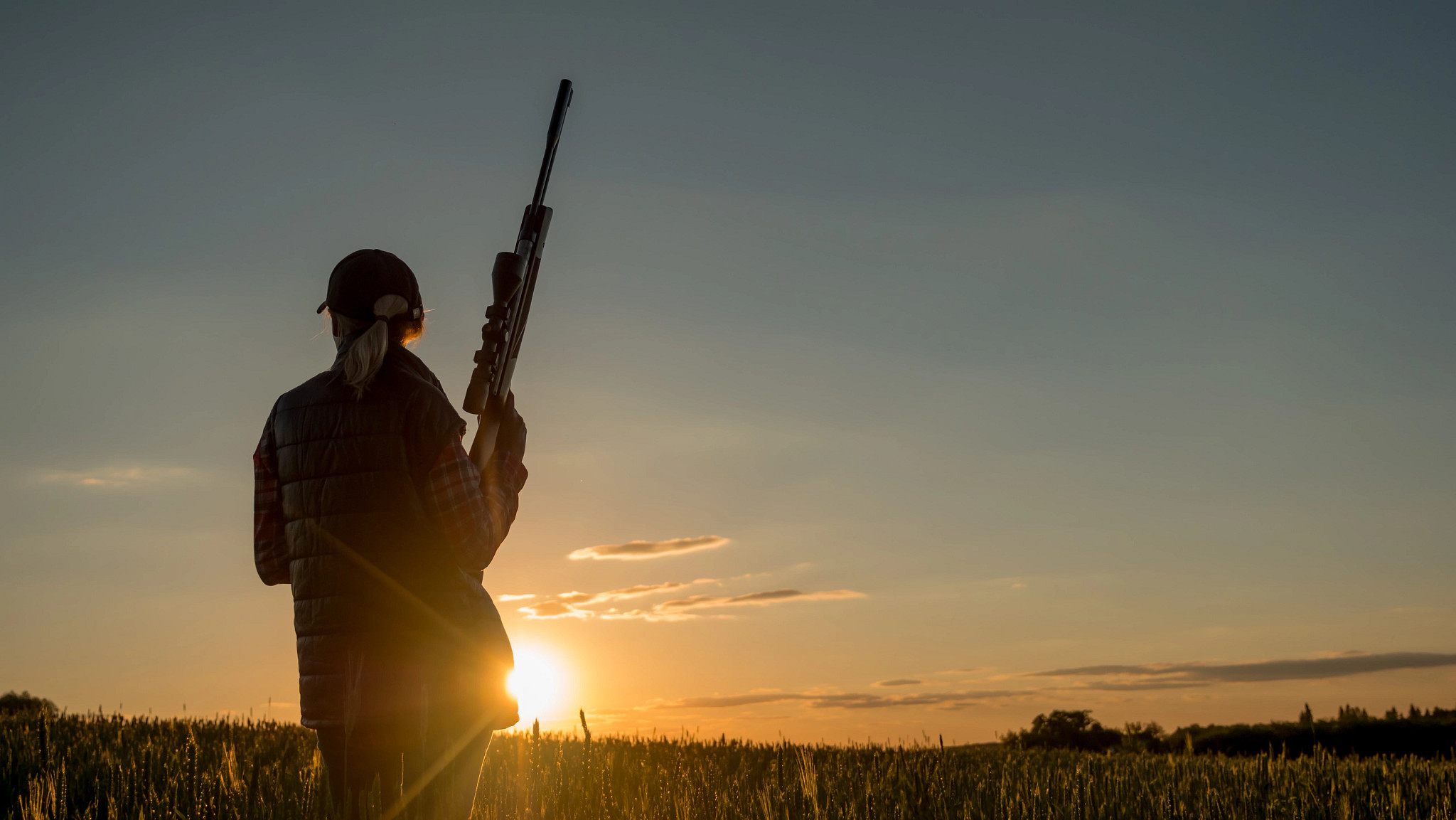 A woman holds a rifle while standing in a field, silhouetted by the rising sun