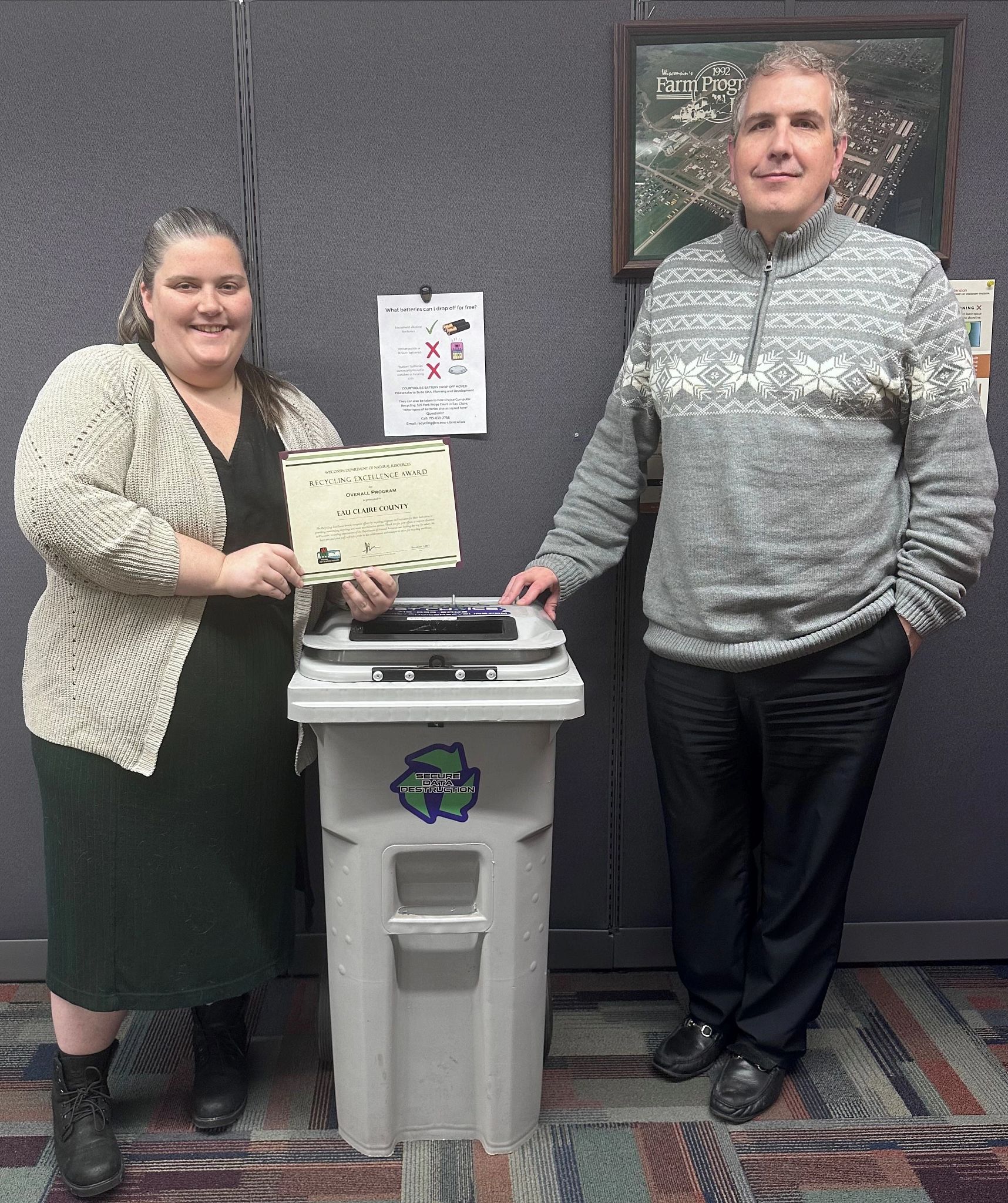 Two team members from Eau Claire County stand next to a white recycle bin and pose with the county's 2023 Wisconsin Recycling Excellence Award certificate from the DNR.