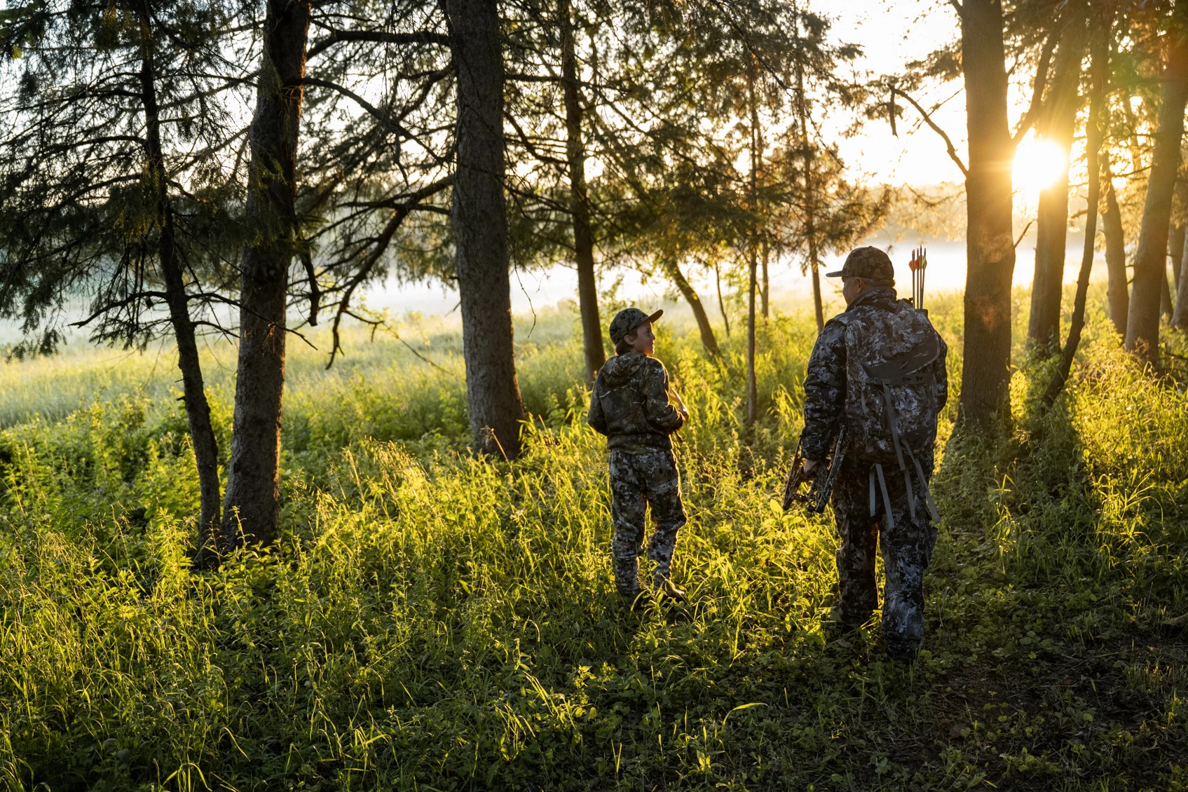 A youth hunter and adult hunter walk in the woods wearing camo. 