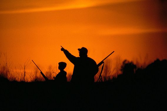 Two hunters standing in a field as the sun sets. One hunter points at something in the sky.