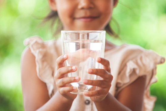 A glass of drinking water is held up to the camera by a girl. 