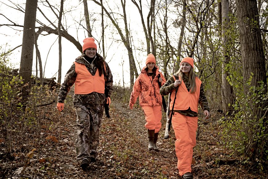 three women hunters wearing camouflage and bright orange walking in the woods