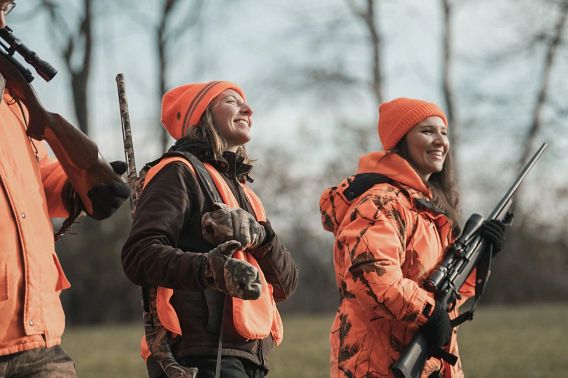 Three women, all wearing blaze orange, walk through a field while hunting.