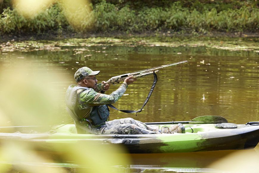 A man in a kayak, wearing camouflage and holding a rifle.
