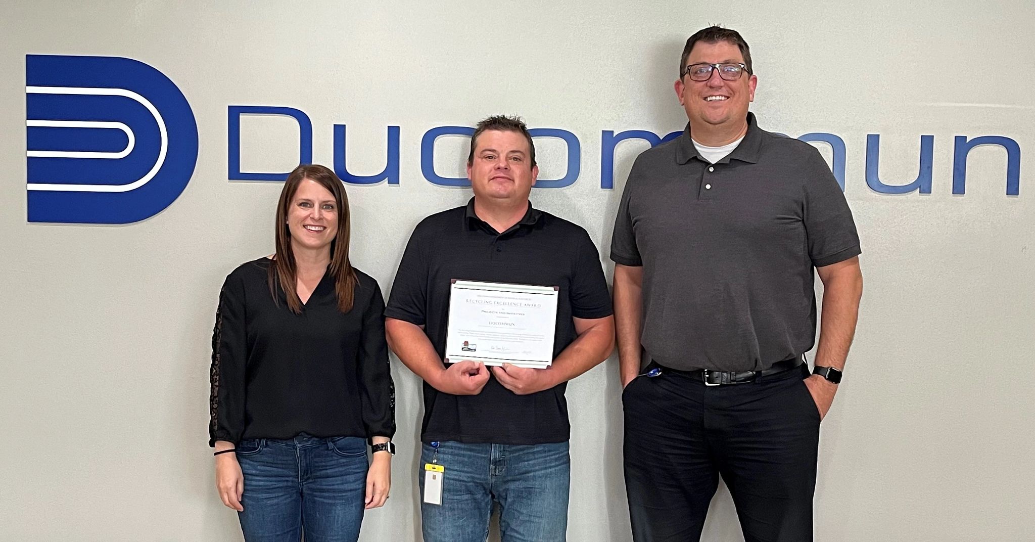 Three employees from Ducommun stand in front of a Ducommon sign while holding their award.