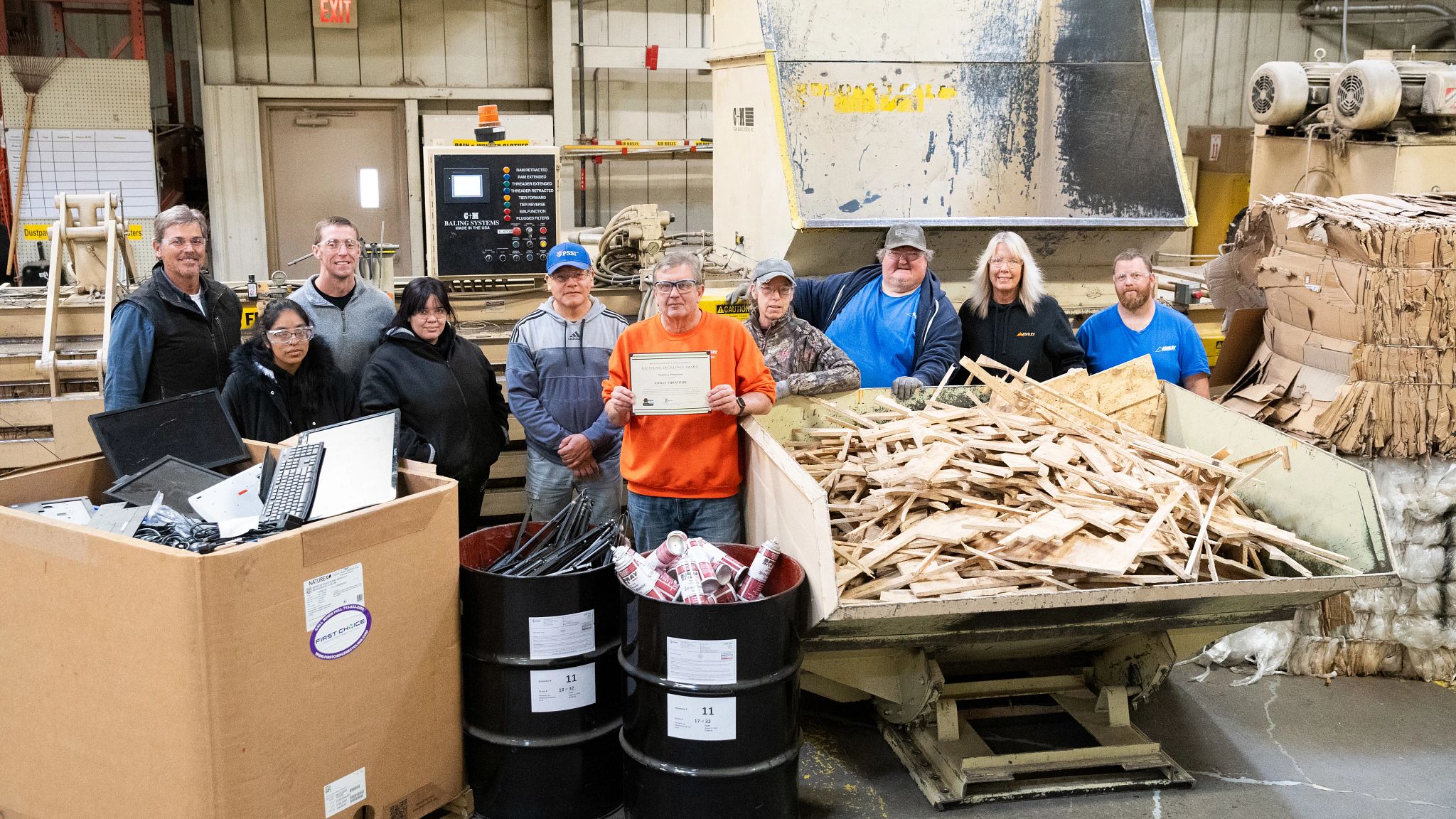 The team at Ashley Furniture gathers around a box, two 50-galloon barrels and a trailer full of recyclable scrap. The team member in the center holds the business's 2023 Wisconsin Recycling Excellence Award certificate from the DNR.