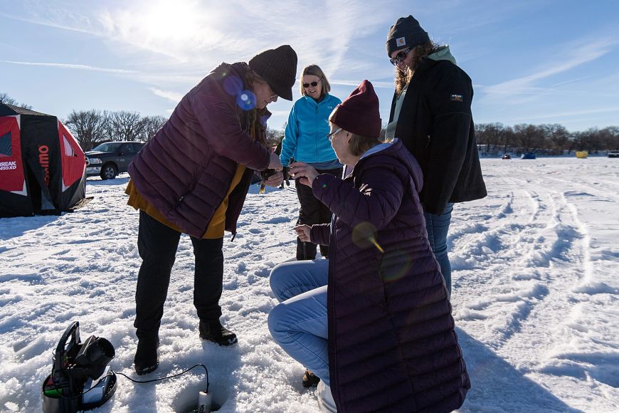 Group of people ice fishing on a snowy landscape with one person crouching by a fishing hole and others standing around in winter clothing.