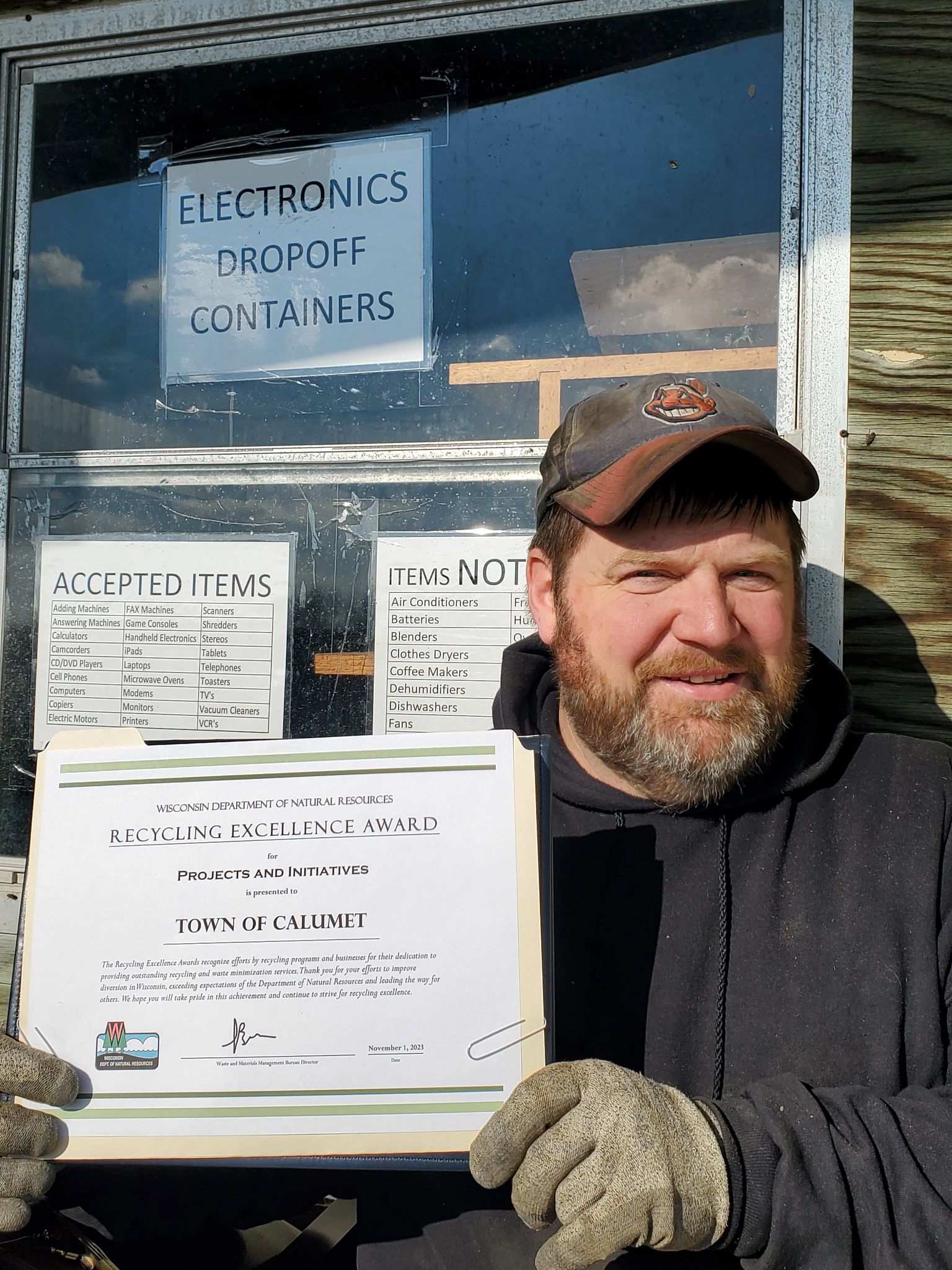 A representative from the Town of Calumet holds the town's 2023 Wisconsin Recycling Excellence Award certificate from the DNR at the town's drop-off site in front of a window listing the acceptable and not-acceptable dropoff items.