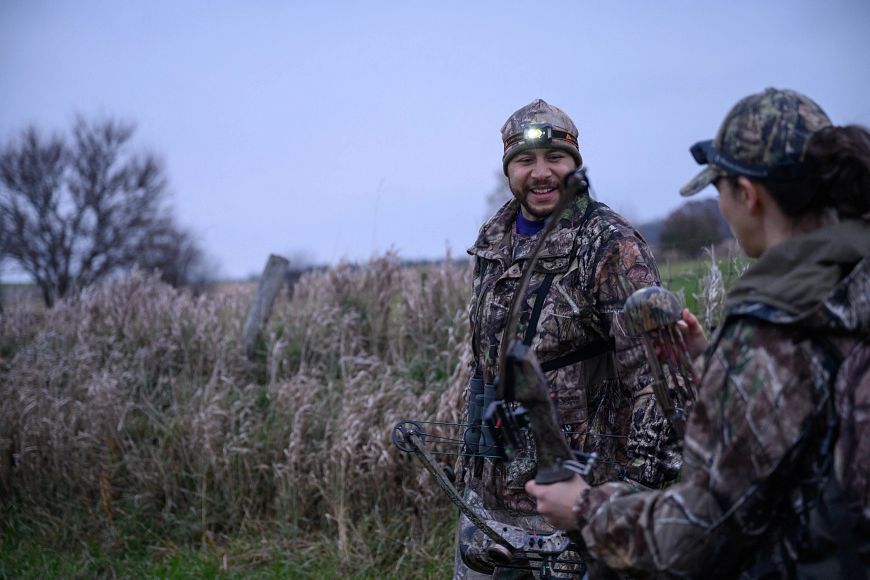 A man and a woman wearing camouflage, walking out in a field to go bow hunting.