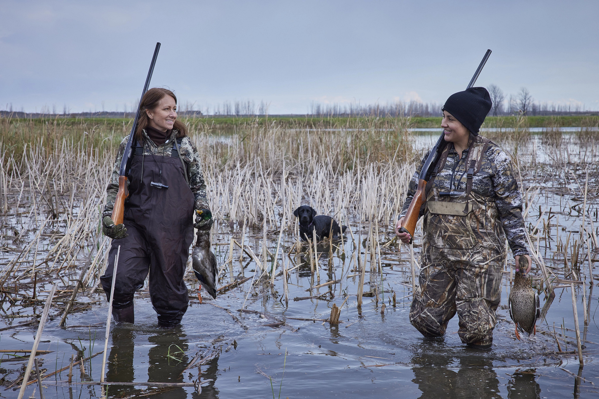 two women wearing camo waders stand in water while duck hunting