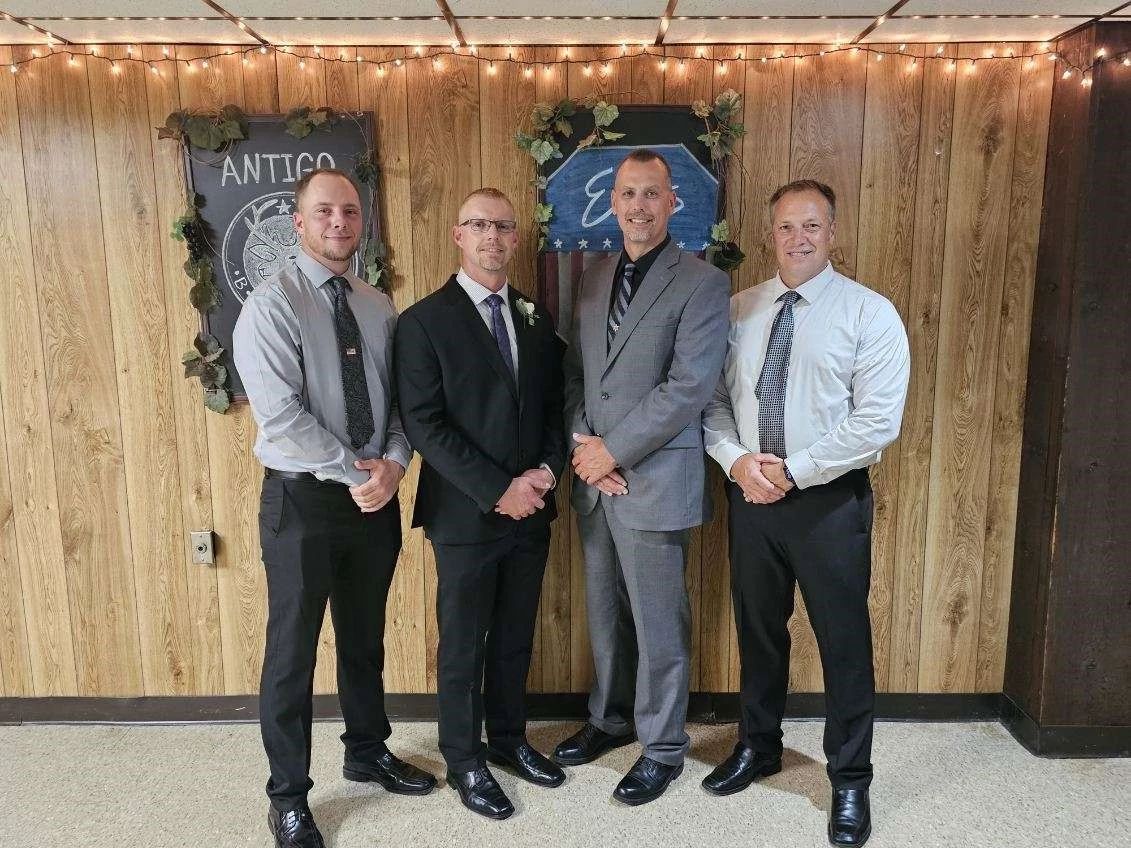 Four individuals stand side by side in formal attire in front of a wood-paneled wall with a decorative emblem and floral arrangements.