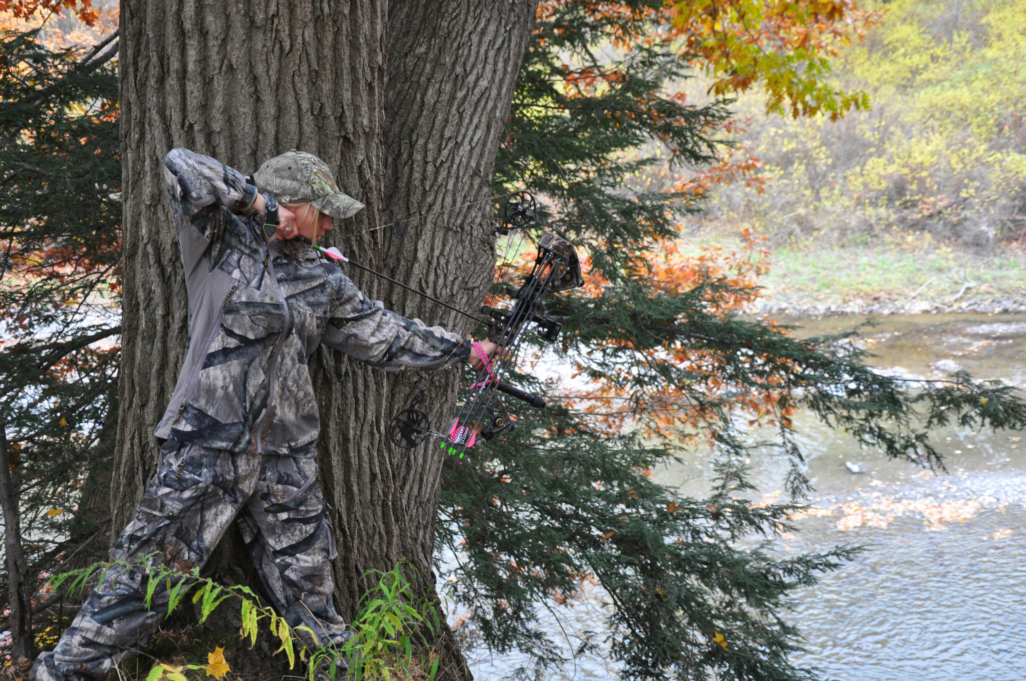 Female hunter with her bow pulled back, leaning against a tree. 