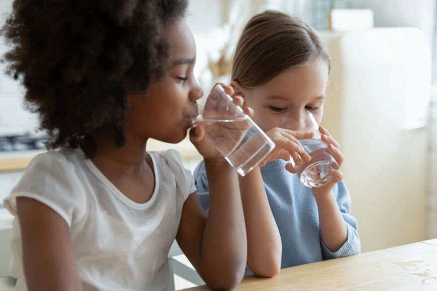 Two little girls sit at table in kitchen drinking water.