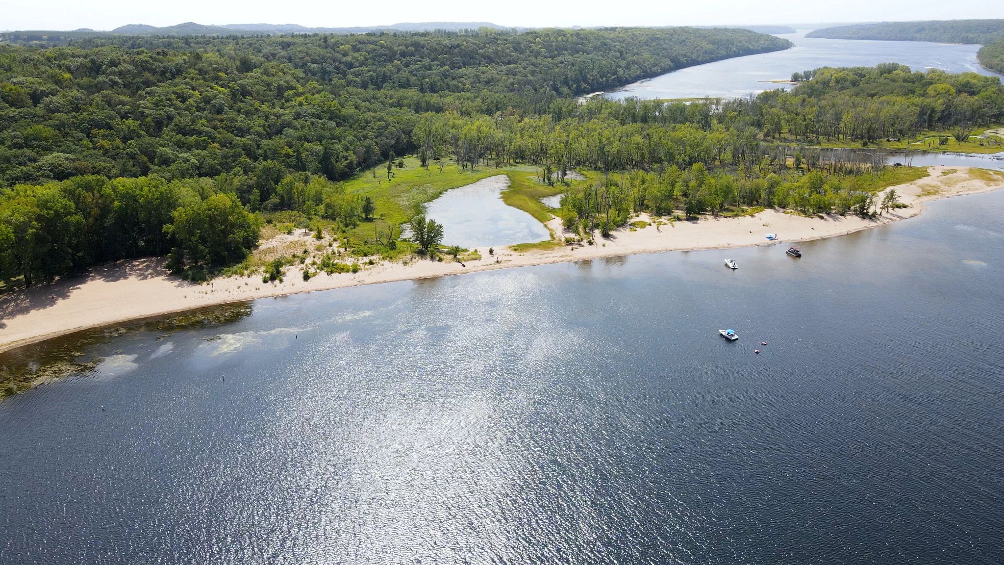Aerial view of a river with a motorboat pulling a person on an inflatable raft, other boats nearby, surrounded by lush greenery and a sandy shore.