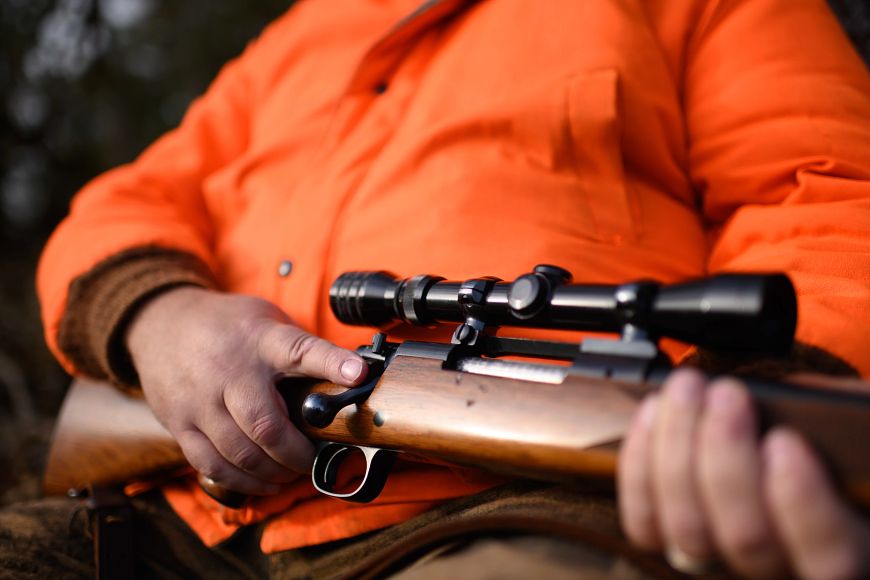 A person in an orange jacket holds a rifle with a wooden stock and a black scope across their lap. 