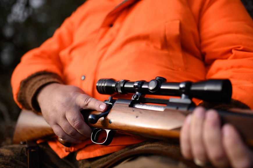 A person in an orange jacket holds a rifle with a wooden stock and a black scope across their lap.