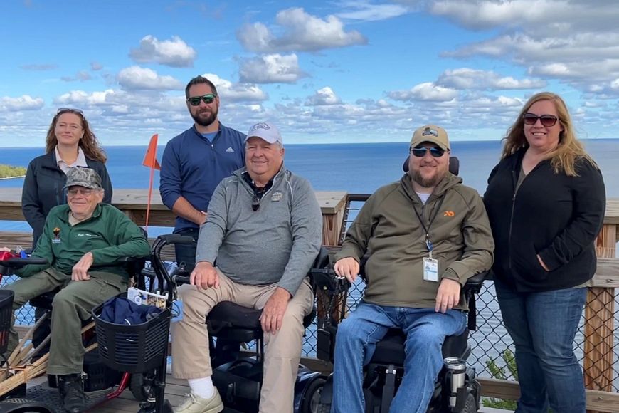 Six people posing for a photo at the top of Eagle Tower at Peninsula State Park with Lake Michigan and fluffy white clouds in the background.