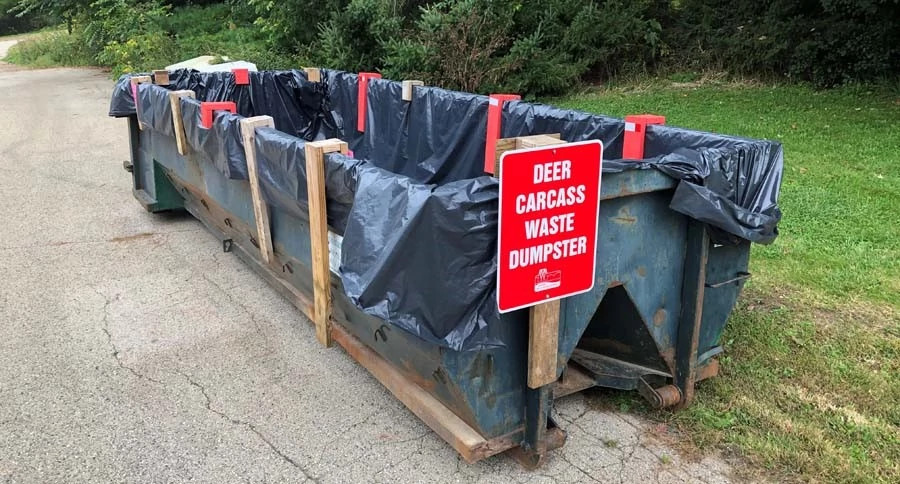 A black deer carcass disposal dumpster with a red "Deer Carcass Waste Dumpster" sign.