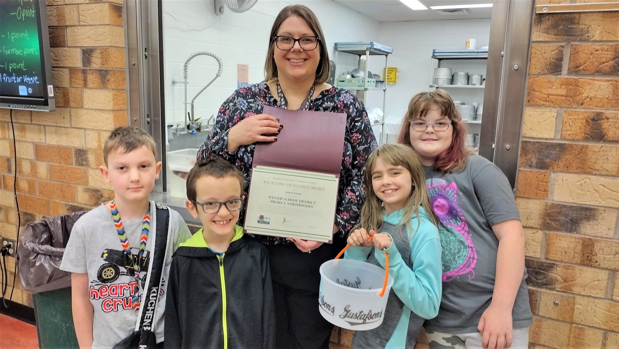 A representative from the Winter School District hold the district's 2023 Wisconsin Recycling Excellence Award certificate from the DNR. She is surrounded by four children from the district, one of whom is holding an empty ice cream bucket that the school uses to collect fruit and vegetable food waste.