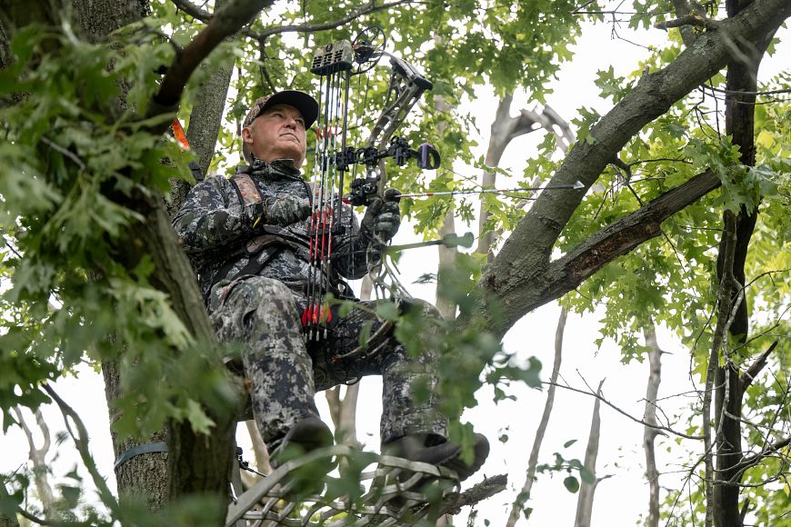 A man in camouflage sits in an elevated tree stand holding a compound bow at the ready.