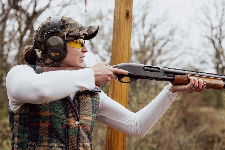 a woman wearing safety gear shooting a shotgun at a shooting range