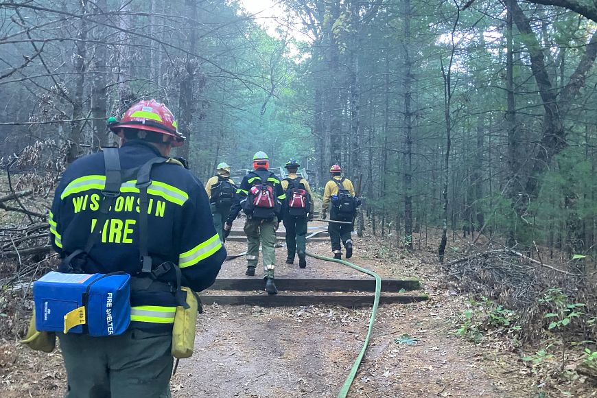 A group of fire fighters, wearing hard hats and carrying axes and other fire fighting gear, walk through a trail in the woods with smoke visible in the air.