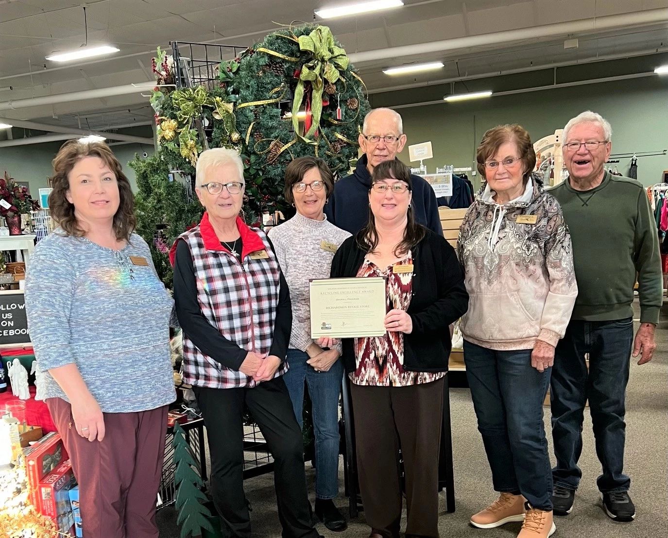 Seven members of the Richardson Resale Store pose in their store in front of a holiday wreath display. The team member in the center holds the business's 2023 Wisconsin Recycling Excellence Award certificate from the DNR.